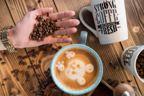 Manos femeninas con granos de café, una taza de café con espuma junto al molinillo de café en la mesa de madera, vista superior — Foto de Stock