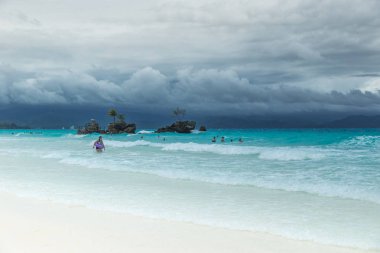 White Beach of Boracay and people swim at turquoise azure Sea on a cloudy day near Grotto Willys Rock upon which stands the Virgin Mary Statue. Boracay, Philippines clipart