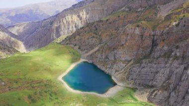 Turkuaz su ve yeşil çimlerle bir dağ gölünün etrafında uçmak. Yansıma gökyüzü ve sudaki bulutlar. Yüksek dağları ve gölü olan güzel bir bahar manzarası. Hava görüntüsü 4K.