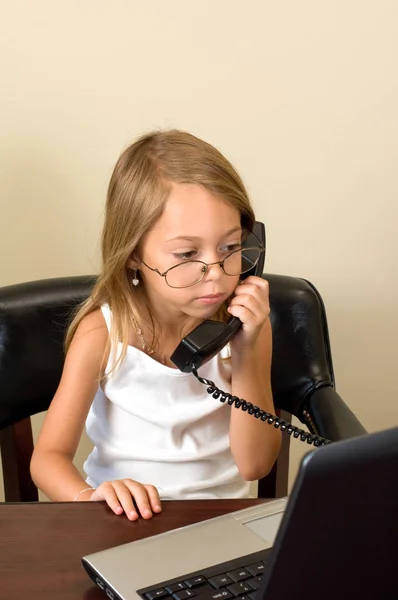 A young girl about 6 pretends to be busy at her notebook computer while wearing glasses too big for her head, talking on a telephone.