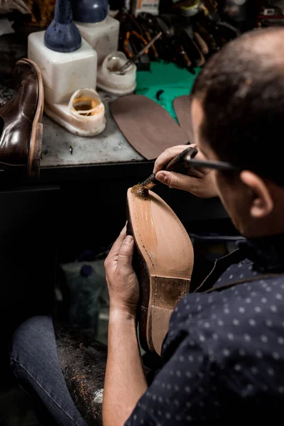 Male cobbler sitting at his shop and repairing man's shoe, brushing glue over sole.