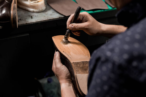Male cobbler sitting at his shop and repairing man's shoe, brushing glue over sole. 