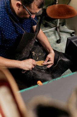 Erkek ayakkabıcı klasik görünümlü atölyesinde oturuyor ve ayakkabı tamiri için deri kumaş kesiyor.