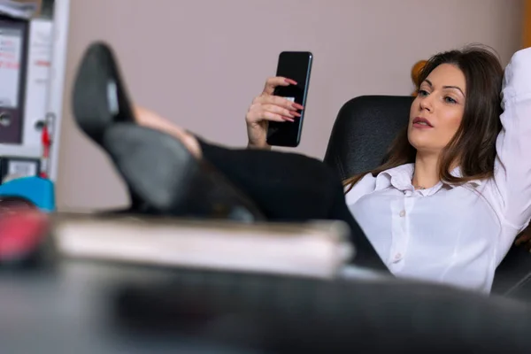 Güzel bir iş kadını ofisinde oturuyor, bacakları masanın üstünde ve akıllı telefonuna bakıyor.
