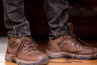 Koyu kotlu bacakları ve tahta zeminde kahverengi deri çizmeleri olan, bağcıksız ayakkabı bağı olan bir adam. 