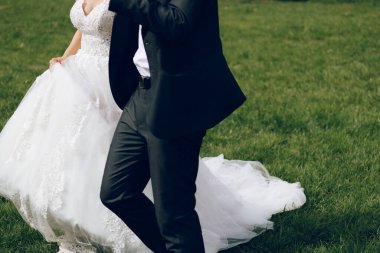 Klasik takım elbiseli damat ve uzun beyaz gelinlikli gelin yeşil çimlerde yürüyor ve el ele tutuşuyor. Tanınabilir yüz yok. 