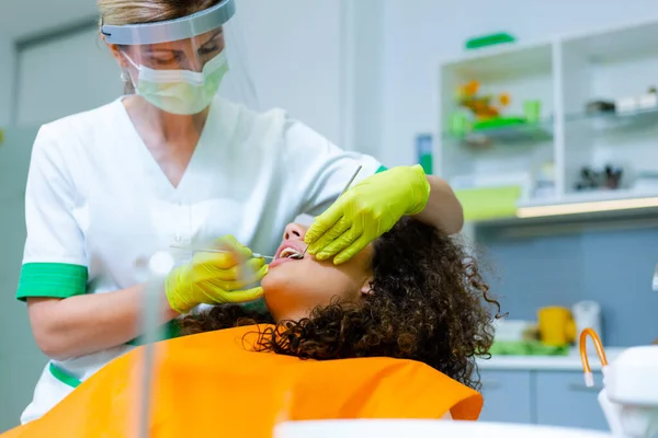  Orta yaşlı beyaz bir kadın tarafından yapılan diş kontrolünde güzel kıvırcık saçlı melez bir genç kız. Coronavirus güvenlik önlemi dişçinin ofisinde. 