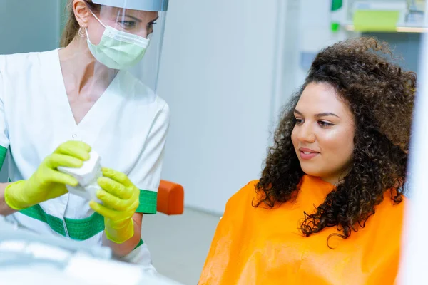 Caucasian female dentist talking to mixed race teenage patient, explaining importance of oral hygiene on jaw model.