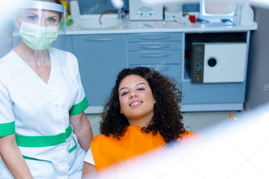 Beautiful mixed-race teenage girl on dental checkup by middle-age caucasian woman wearing face mask and visor as Coronavirus safety precaution. Modern dentist's office. 