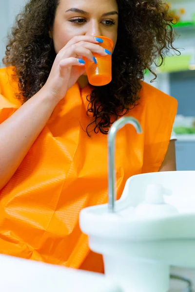 Teenage girl rinse mouth at dental office, drinking water from plastic orange color cup.