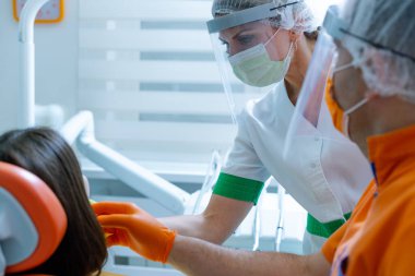 Kadın dişçi ve erkek teknisyen hasta ile görüşüyor. Diş hekimi maske, güneşlik ve eldiven takıyor. Koronavirüs güvenlik önlemi olarak.. 