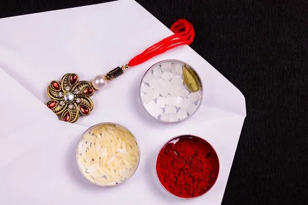 Hint Festivali - Pilav taneleri, kumkum, şeker kayası ve kakule kaplı zarif Rakhi beyaz bir zarfın içinde, siyah arka planda. Raksha Bandhan konsepti