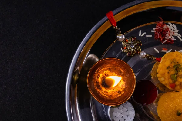 Indian Festival of Brothers and Sisters - Rice grains, kumkum, sweets and diya on plate with an elegant Rakhi on balck background. A traditional Indian wrist band which is a symbol of love