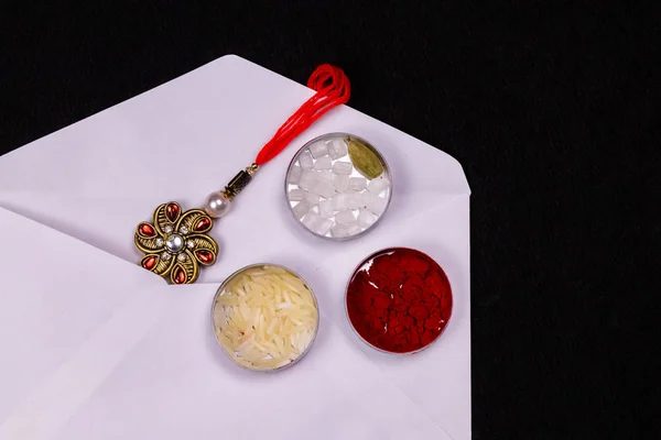 Raksha Bandhan kavramı - Siyah arka planda düz bir teneke kutuda pirinç taneleri, kumkum ve şeker içeren beyaz bir zarfta Zarif bir Rakhi