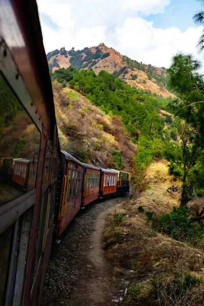 tren yan taraftaki ağaçlar ve dağlar boyunca ilerliyor. Gökyüzü bulutlu.
