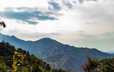 Geniş bir dağ sırası bulutlu gökyüzü ve ön planda ağaçlarla.