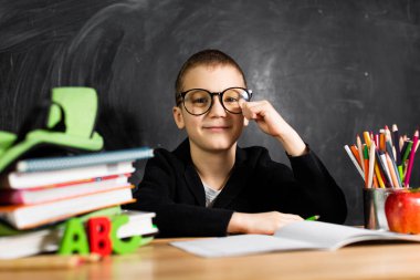 Bir okul çocuğu kameraya gülümseyen bir masada oturur, yine okulda, eğitimdedir..
