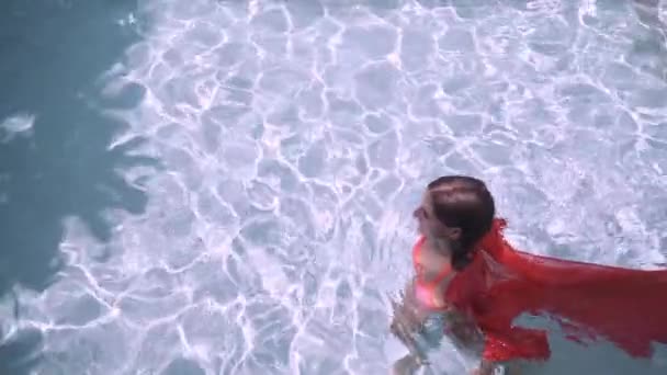 Bella ragazza in un abito lungo rosa nuota in piscina. Affascinante bruna in un abito lungo rosa posa per la fotocamera in una piscina blu . — Video Stock