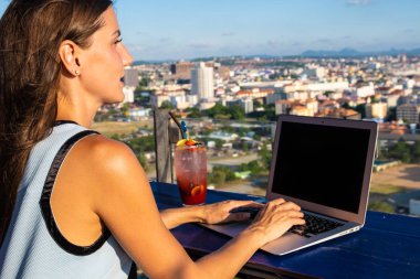 Kız çatıdaki bir kafede bilgisayarda çalışıyor. Çatı katındaki kafede dizüstü bilgisayara yazı yazan kadın, şehrin panoramik manzarasına sahip ahşap bir masada oturuyor..