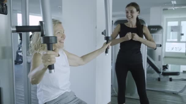 Pattaya, thailand - 15. Mai 2019: Eine Seniorin sitzt mit einem Personal Trainer an einem Simulator im Fitnessstudio. Tochter hilft Mutter in der Turnhalle — Stockvideo