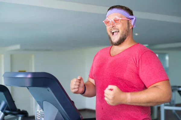 Pembe bir T-shirt ve pembe gözlük içinde oynak şişman adam spor salonunda fitness meşgul.