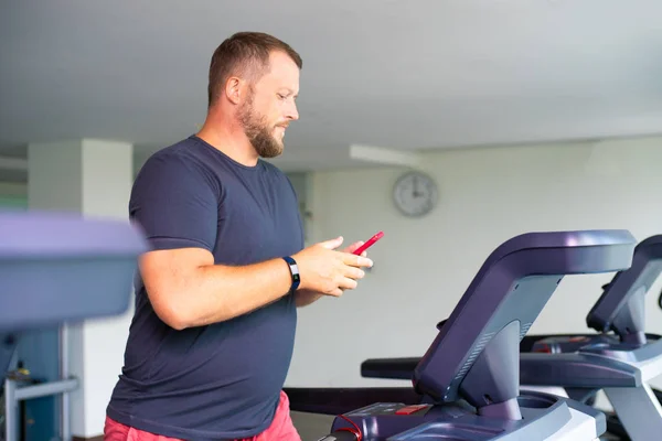 Hombre regordete caminando en la pista, calentando en la cinta de correr del gimnasio. hombre con teléfono — Foto de Stock