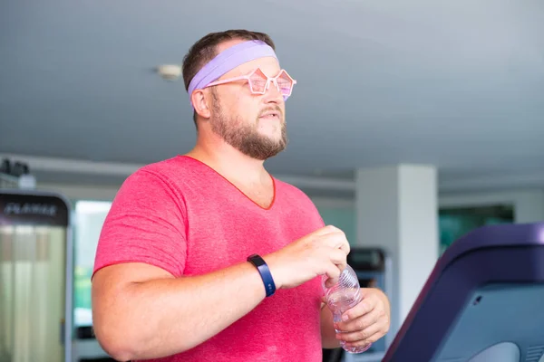 Playful fat man in a pink T-shirt and pink glasses is engaged in fitness in the gym. man drinking water in the gym