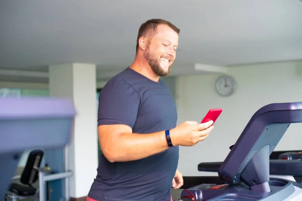 chubby man walking on running track, warming up on gym treadmill. man with phone