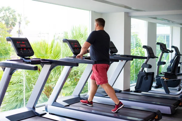 chubby man walking on running track, warming up on gym treadmill. back view