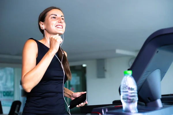 Mujer en forma joven corriendo en la cinta de correr escuchando música a través de auriculares en el gimnasio — Foto de Stock