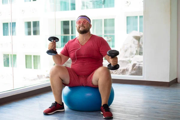 Playful fat man in a pink T-shirt and pink glasses is engaged in fitness with dumbbells and a fit ball in the gym. man drinking water in the gym. man listening to music on headphones