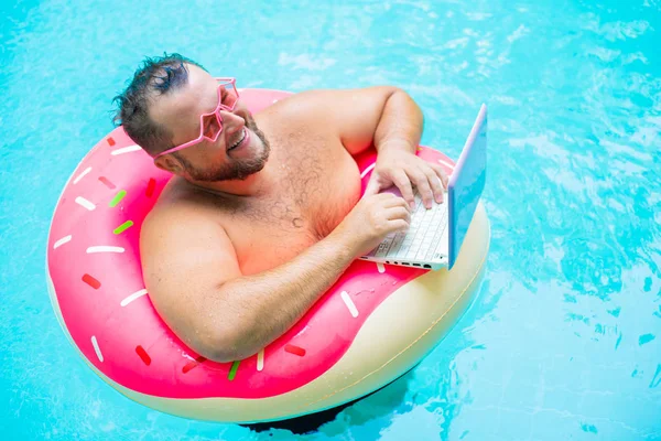Lachende grappige dikke man in roze bril op een opblaasbare cirkel in het zwembad werkt op een laptop die een meisje portretend. — Stockfoto