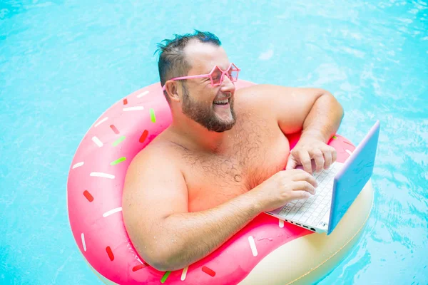 Lachende grappige dikke man in roze bril op een opblaasbare cirkel in het zwembad werkt op een laptop die een meisje portretend. — Stockfoto