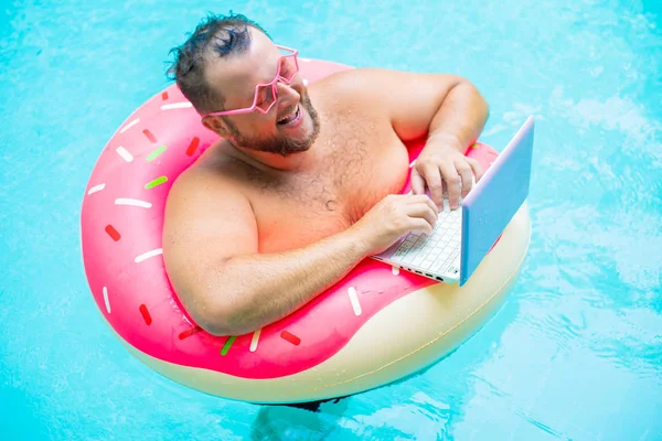 Lachende grappige dikke man in roze bril op een opblaasbare cirkel in het zwembad werkt op een laptop die een meisje portretend. — Stockfoto