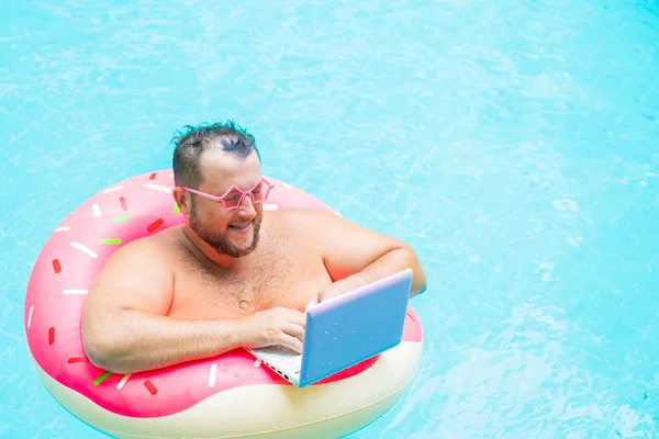 Enjoying Funny fat male in pink glasses on an inflatable circle in the pool works on a laptop portraying a girl. — Stock Photo, Image