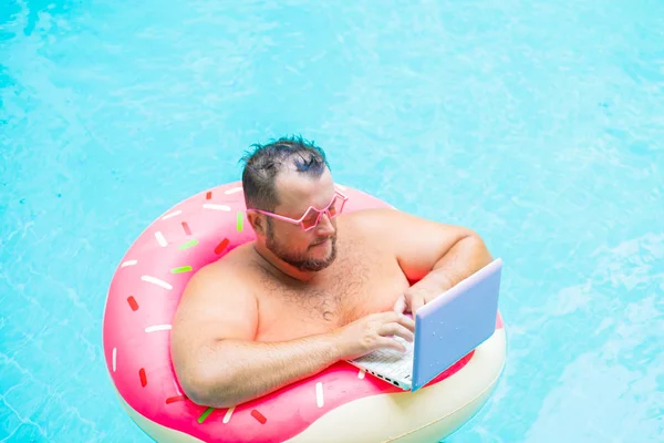 serious Funny fat male in pink glasses on an inflatable circle in the pool works on a laptop portraying a girl.