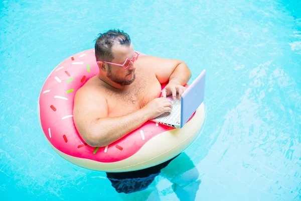 serious Funny fat male in pink glasses on an inflatable circle in the pool works on a laptop portraying a girl.