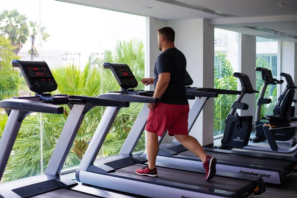 chubby man walking on running track, warming up on gym treadmill. back view