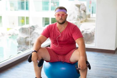 Pembe t-shirt ve pembe gözlük lütfedilen şişman adam halterve spor salonunda uygun bir top ile fitness yapan. Spor salonunda su içen adam. kulaklıkla müzik dinleyen adam