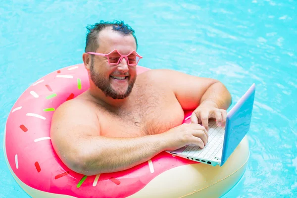 Enjoying Funny fat male in pink glasses on an inflatable circle in the pool works on a laptop portraying a girl. — Stock Photo, Image