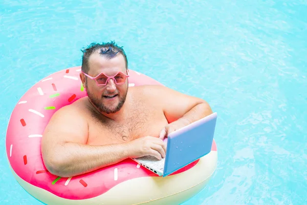 Verraste grappige dikke man in roze bril op een opblaasbare cirkel in het zwembad werkt op een laptop die een meisje portretend. — Stockfoto