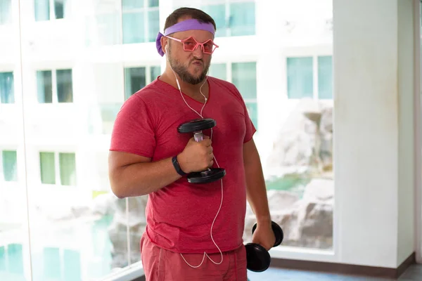 Playful fat man in a pink T-shirt and pink glasses is engaged in fitness in the gym. man drinking water in the gym. man listening to music on headphones