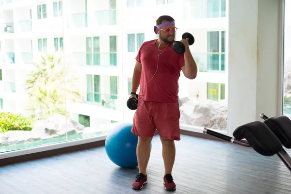Playful fat man in a pink T-shirt and pink glasses is engaged in fitness with dumbbells and a fit ball in the gym. man drinking water in the gym. man listening to music on headphones