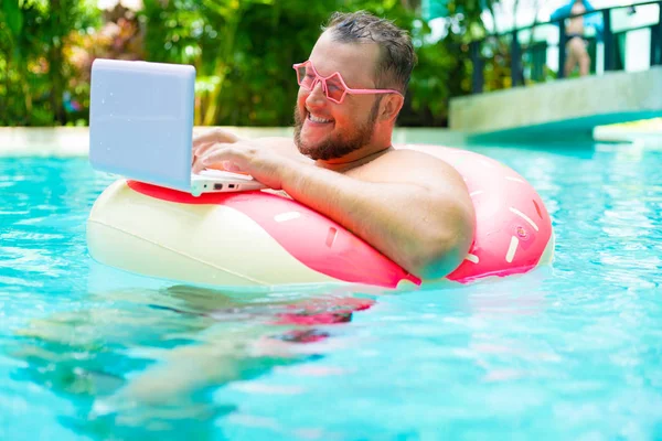 Funny fat male in pink glasses on an inflatable circle in the pool works on a laptop portraying a girl. — Stock Photo, Image
