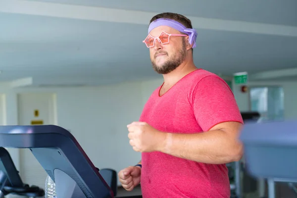 Playful fat man in a pink T-shirt and pink glasses is engaged in fitness in the gym.