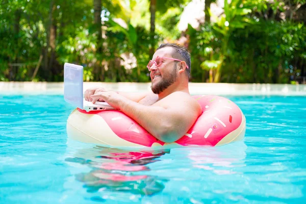 Lachende grappige dikke man in roze bril op een opblaasbare cirkel in het zwembad werkt op een laptop die een meisje portretend. — Stockfoto