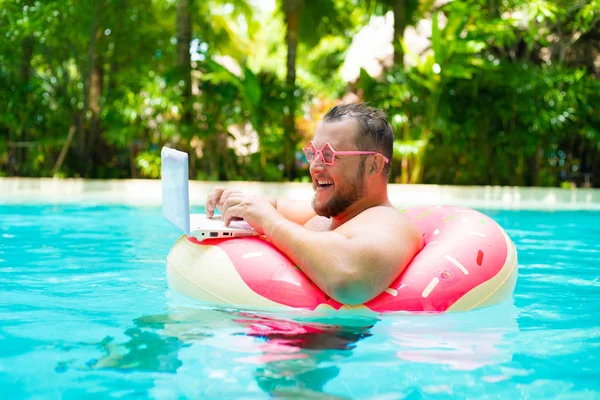 Funny fat male in pink glasses on an inflatable circle in the pool works on a laptop portraying a girl.