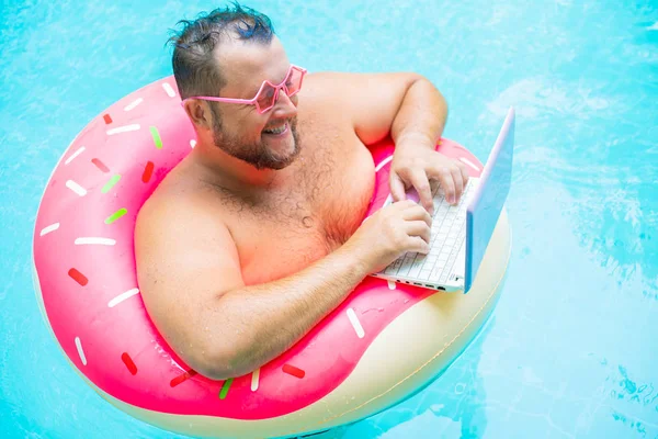 enjoying Funny fat male in pink glasses on an inflatable circle in the pool works on a laptop portraying a girl.