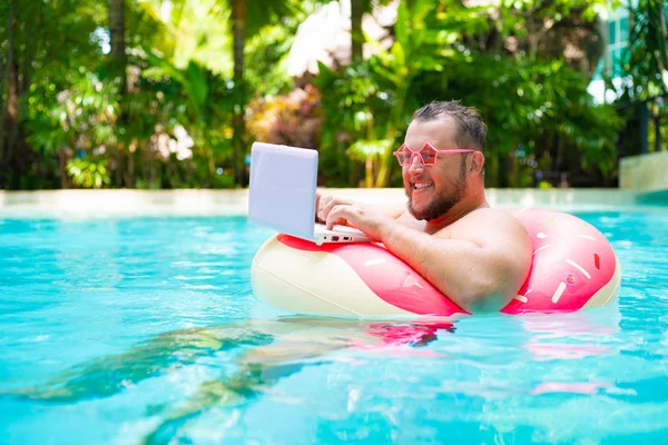 Funny fat male in pink glasses on an inflatable circle in the pool works on a laptop portraying a girl.