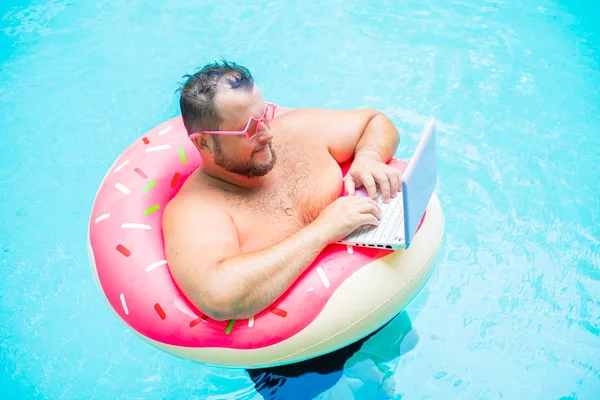 Ernstige grappige dikke man in roze bril op een opblaasbare cirkel in het zwembad werkt op een laptop portretend een meisje. — Stockfoto